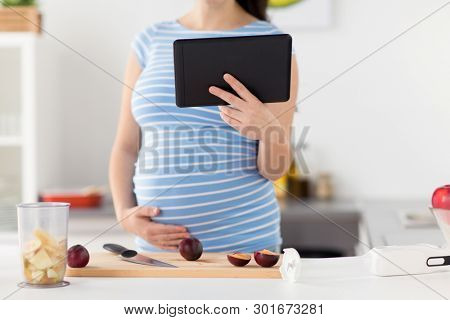 cooking, pregnancy and technology concept - pregnant woman with tablet computer, kitchen knife and plums on wooden cutting board at home