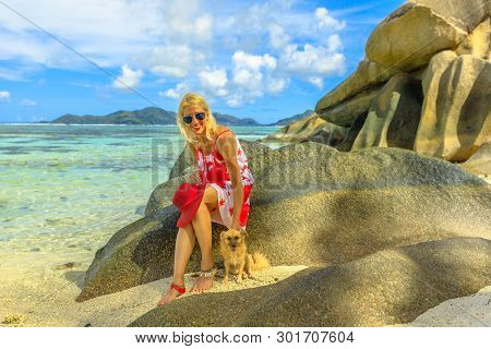 Smiling Lifestyle Woman Sitting On A Boulders, Touches A Cute Dog At Anse Source Dargent One Of The 