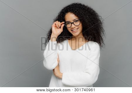 Portrait Of Happy Woman With Curly Hair, Kepes Hands On Frame Of Glasses, Has Gentle Smile, Wears Wh