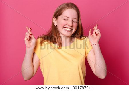 Cheerful Young Beautiful Girl With Crossed Fingers, Makes Wish, Smiles Broadly, Wears Casual Yellow 