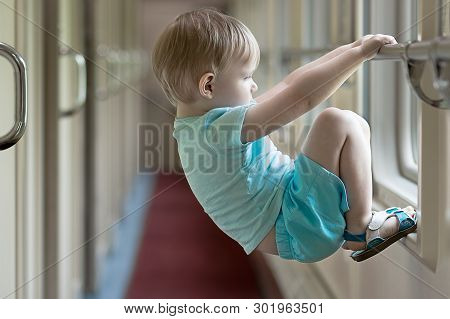 A Bored And Sad Blond Boy Toddler Hangs On The Crossbar Of A Train Window. Concept For Articles Abou