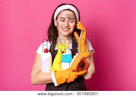 Positive Caucasian Woman Speaking On Mobile Phone While Clening Her House, Holding Sponge And Deterg