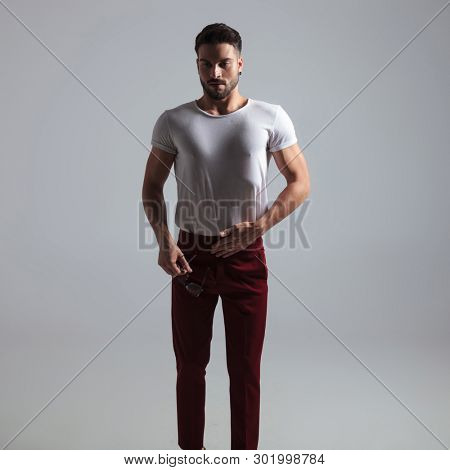 Thoughtful guy holding his breath and his pair of glasses while wearing a white t-shirt and red pants, standing on white studio background