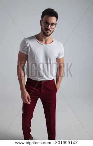 Charming man romantically looking to the camera and holding his hand in his pocket while wearing glasses, a white t-shirt and red pants, standing on gray studio background