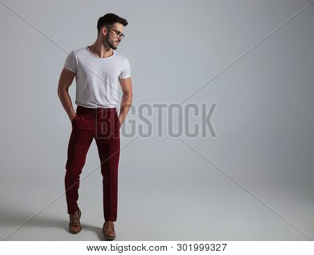 Casual guy looking to the side and holding both of his hands on his pockets while wearing glasses and a white t-shirt, standing on white studio background