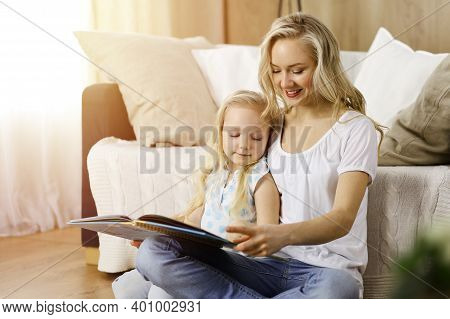 Happy Family. Blonde Young Mother Reading A Book To Her Cute Daughter While Sitting At Wooden Floor 