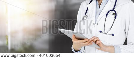 Unknown Woman Doctor Iusing Tablet Computer In Her Hands, While Standing In Sunny Clinic. Medicine C