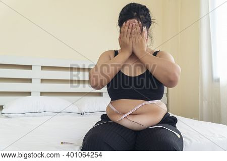 Obesity Unhealthy Concept. Overweight Asian Woman Sitting On White Bed While Holding Hands Cover On 