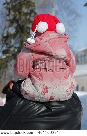 A Small Child Sits On His Father's Shoulders. On The Child's Head Is A Red Hat With White Pampons. P