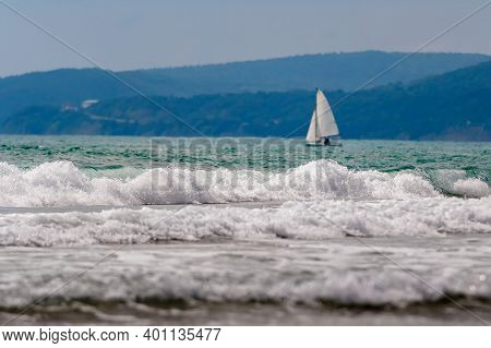 Seascape, Small Waves With White Foam At The Top Against The Background Of Blue Water. A Lone Sailbo