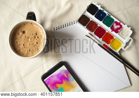 Set Of Watercolor Paints, Scetchbook, Paintbrush And A Cup Of Coffee For Breakfast.