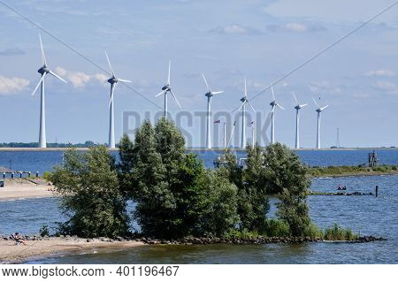 Urk, The Netherlands - June 15 2020 Windmills On The Coast With People Sunbathing And Fishing, Trees