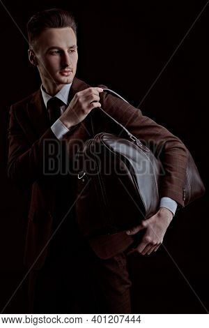 Portrait of a handsome man in elegant classic suit and bow tie posing with leather bag on a black background. Business style. Men's fashion, accessories.