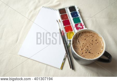 Set Of Watercolor Paints, Scetchbook, Paintbrush And A Cup Of Coffee For Breakfast.