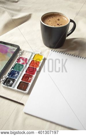 Set Of Watercolor Paints, Scetchbook, Paintbrush And A Cup Of Coffee For Breakfast.