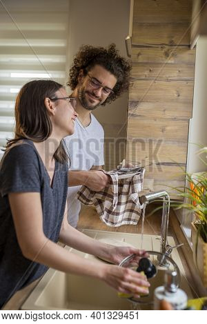 Young Couple In Love Doing The Washing Up And Drying Together After Lunch; Woman Washing While Man I