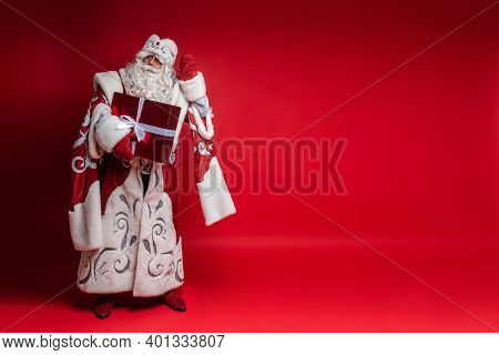 Santa Claus With Xmas Gift Puts His Hand To Head, Tryes To Listen On Red Background With Copy Space 