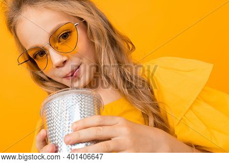 Portrait Of Young Caucaian Girl With Long Fair Hair In Yellow Glasses Drinks A Juice And Smiles