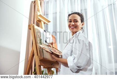 A Smiling Woman Painter Holding A Palette With Paint And A Paintbrush And Standing In A Front Of An 