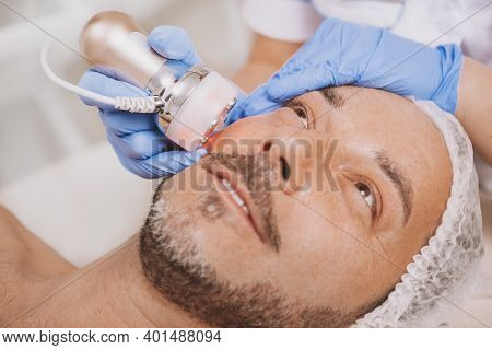 Happy Handsome Mature Man Receiving Facial Hardware Cosmetology Treatment At Beauty Salon. Relaxed A