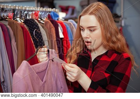 Young Woman Looking Shocked, Checking The Price Of A Dress At Fashion Boutique. Overwhelmed Woman Lo