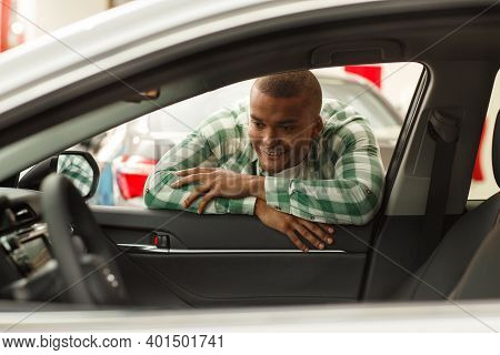 Handsome African Man Examining New Car On Sale Looking Inside The Vehicle. Male Customer Smiling, Ch