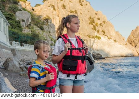 Two Children In Wearing Red Life Jackets Standing On The Beach Near The Rocky Seashore. Equipment Fo
