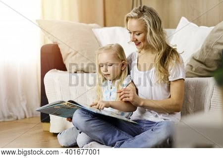 Happy Family. Blonde Young Mother Reading A Book To Her Cute Daughter While Sitting At Wooden Floor 