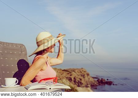 Mature Tourist Woman Relaxing On Coast With Book And Coffee Cup, Enjoy Sea View From Cliff. Reading 