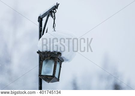 In Winter, An Iron Lantern Hangs On A Pole In The Park. At The Top Of The Lantern Is A Huge Snow Cap