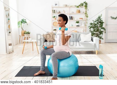 Pregnant Black Woman Sitting On Fitness Ball, Working Out With Dumbbells In Her Home Gym. Expectant 