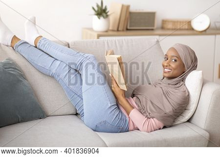 Weekend Stay Home Pastimes Concept. Lovely African American Woman In Hijab Lying On Sofa With Exciti