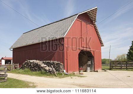 A red storage barn with an opened barn door on a bright, sunny day.  Stacks of wood leans against one side, a distant barn and rail fences on all sides, near and far.