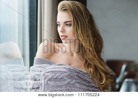 Portrait of seductive blonde young female with long hair wrapped in grey knitted coverlet