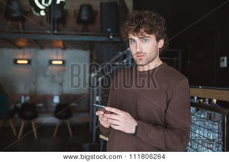 Pensive curly young daydreaming thoughtful handsome man holding mobile phone and using it in cafe