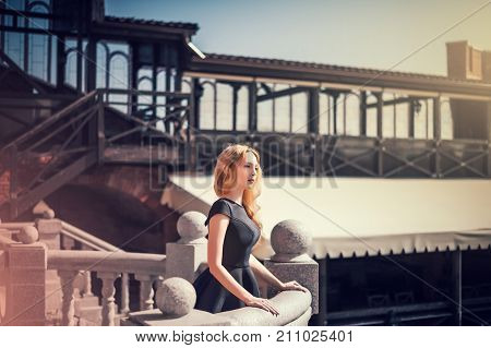 beautiful young blond woman in a black dress and with a fashionable handbag standing on a stone balcony outside