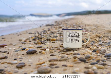 Enameled camp mug on the pebbles of the sandy shore of Lake Baikal during a wave on a mountain background in the summer on a clear sunny day.