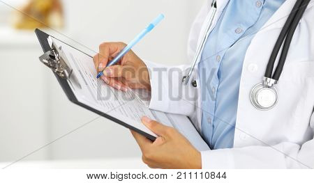 Close-up of a female doctor filling  up medical form at clipboard while standing straight in hospital.