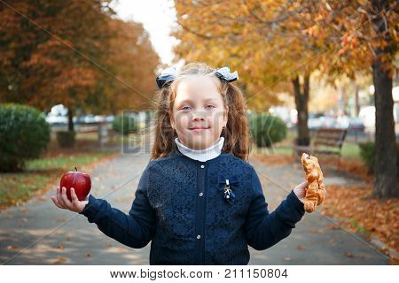 The girl is standing in the park in autumn with an apple and a croissant