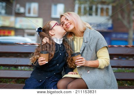 Mom blond in gray sweater with daughter in blue coat on bench in park in autumn daughter kisses mom