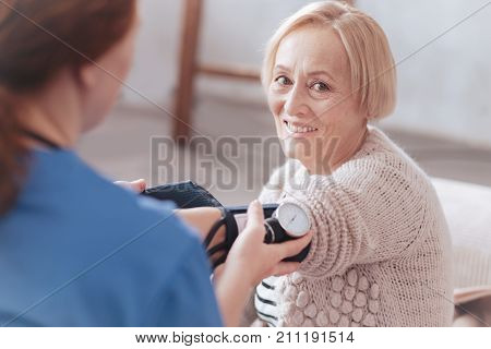 Health care routine. Charming senior woman looking into the camera with a cheerful smile on her face while sitting next to a female doctor and measuring her