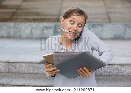 Portrait of crazy young Caucasian businesswoman holding pen in teeth and with laptop and documents in hands talking on mobile phone. Overworking and multi-tasked manager concept