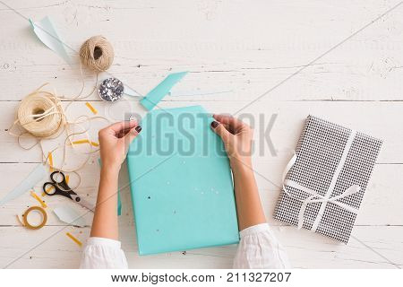 Closeup of woman's hands with gift ribbon twine tape and present wrapped in turquoise gift paper on white wooden background. Christmas birthday or any other celebration preparations.