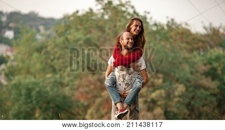 Enamored couple have fun on rest in forest. Young man lifted up girl on his back and carries her. They laugh cheerfully. Closeup.