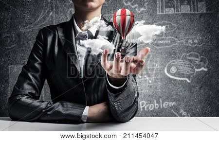 Closeup of business woman in suit presenting flying aerostate among clouds in her palm with business sketches on background. 