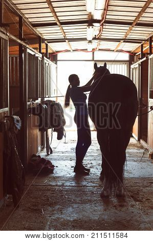 Silhouette of young horsewomen owner harnessing the stallion in stable. Vertical image with vintage filter.