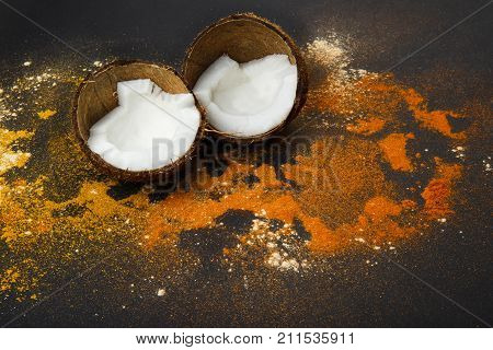 On a black background a split coconut around which spice curry and paprika are scattered. Close up.