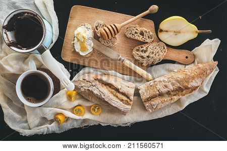 Breakfast set. Cerve pot of coffee, a cup on a kitchen towel, kumquats, pears, baguette slices with butter cream and honey on a rustic wooden board over a black backdrop. Top view