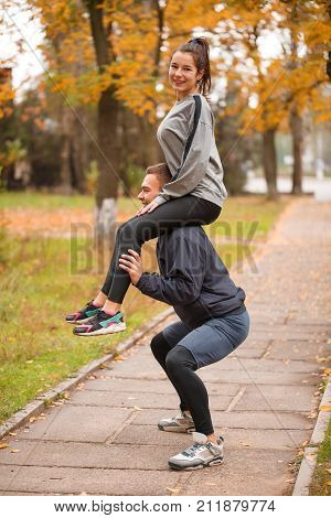 A beautiful young and loving couple are sporting in a park in the fall. The guy crouches holding the girl on his shoulders.
