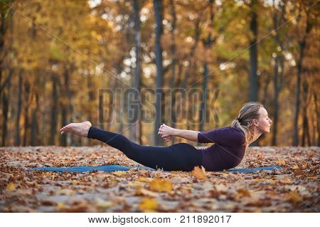 Beautiful young woman practices yoga asana Salabhasana locust pose on the wooden deck in the autumn park.
